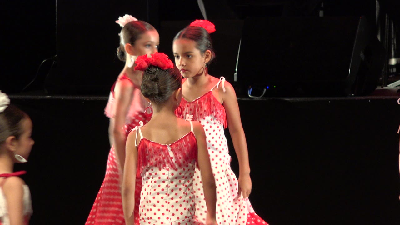 Several girls in red and white dresses remain grouped on the dark Cabo San Lucas stage, with a front-facing child near center-right and two others seen from the back or side.