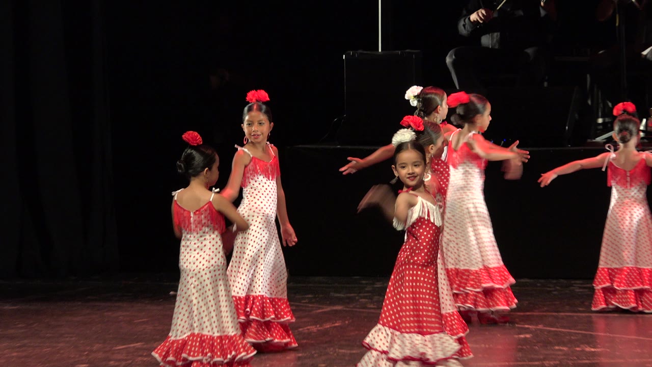 Young girls in red and white dresses dancing on a dark stage.