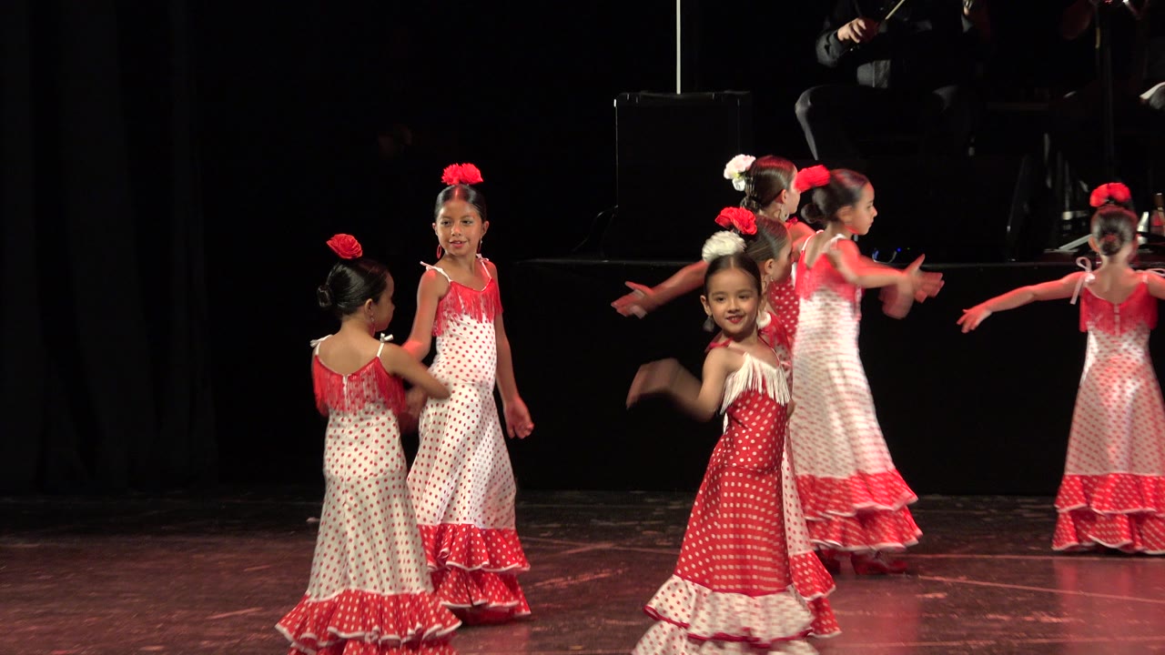 Children in matching red and white dresses continue the dance in Cabo San Lucas, Mexico, with one girl facing the camera while others turn across the stage.