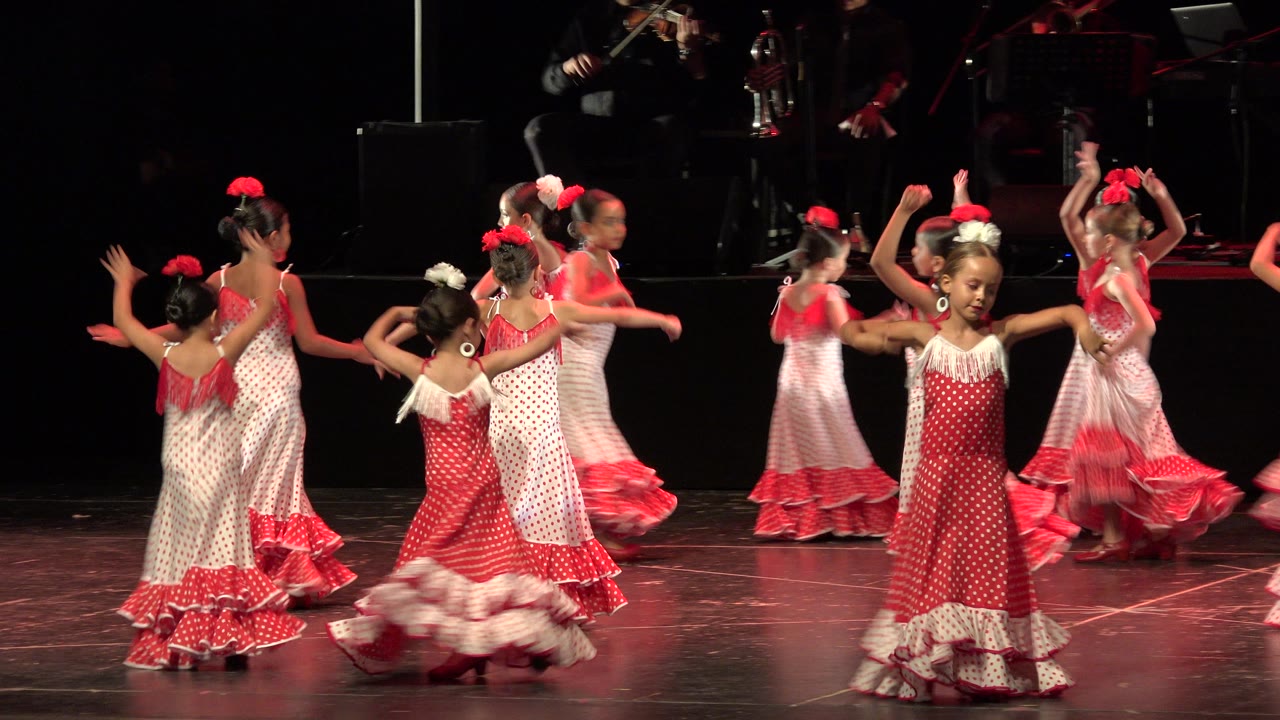At 0:01, several girls turn in place while a front dancer in a red polka-dot dress faces the audience on the Cabo San Lucas stage.