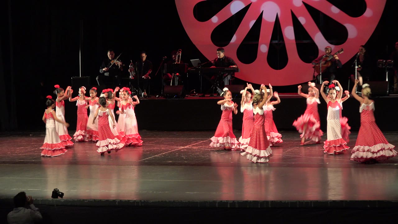 Girls in pink dresses dancing on a theater stage with musicians behind them.