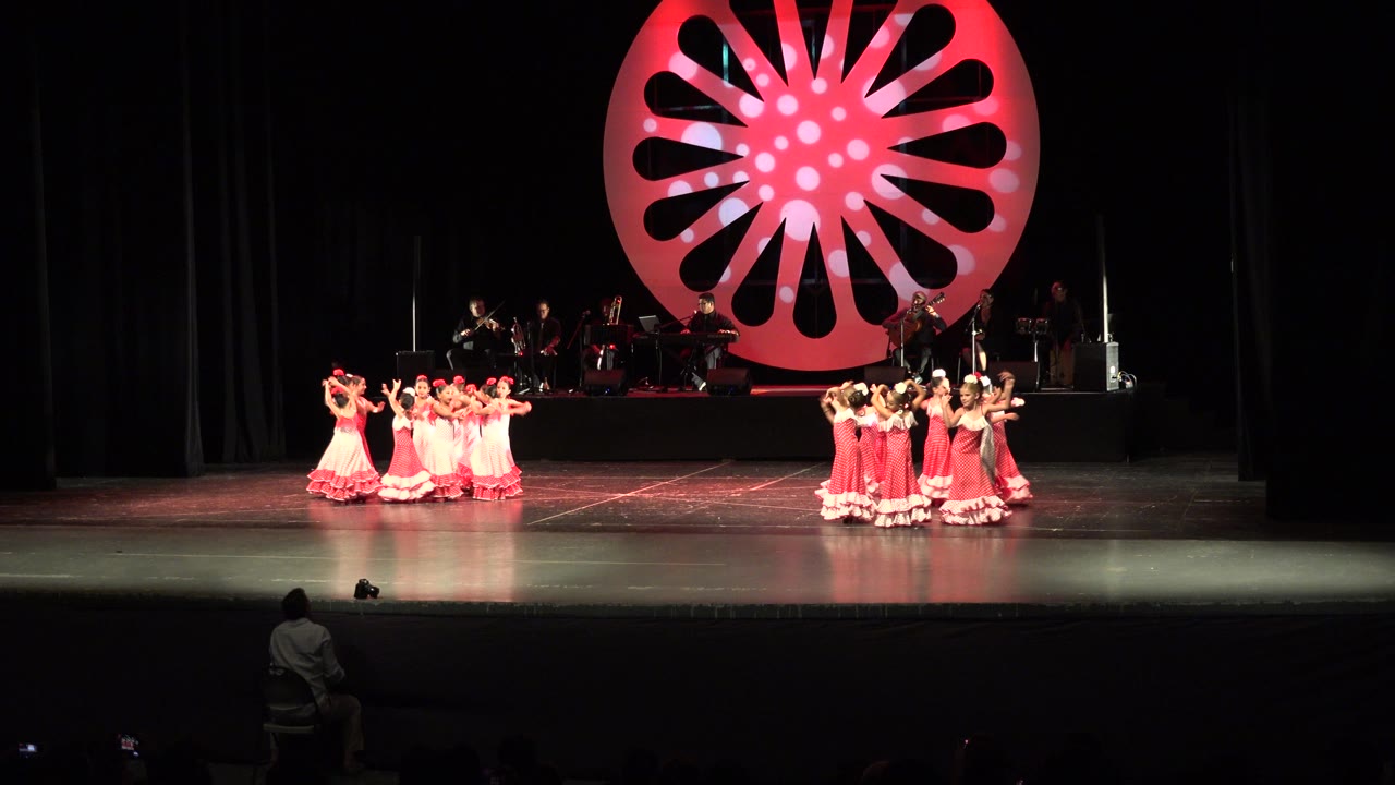 Two groups of young girls in red and pink dresses perform on a dark stage in Cabo San Lucas, Mexico, with musicians behind a large red circular backdrop.