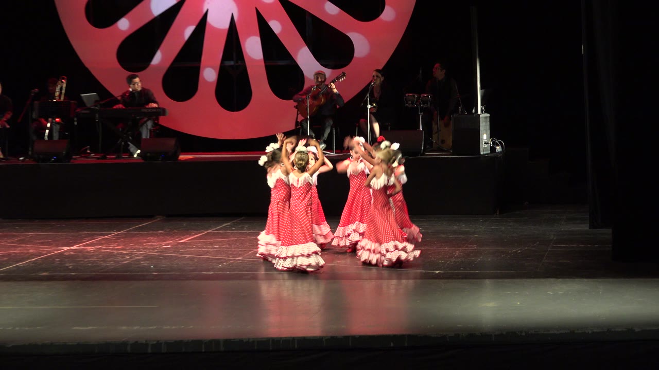 A group of preschool-age girls in red ruffled dresses dances together on a dark stage in Cabo San Lucas, Mexico, with a keyboard player, guitarist, and large red backdrop visible behind them.