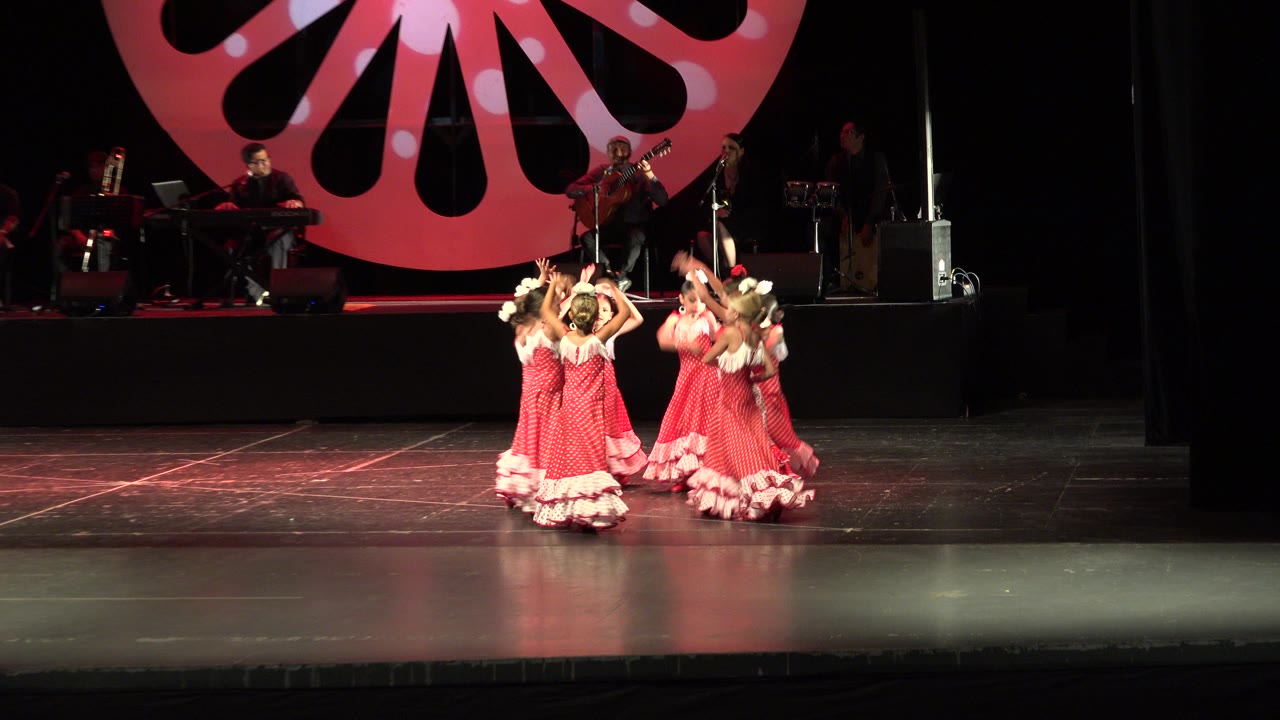 Children in matching red costumes raise their arms while dancing on stage.