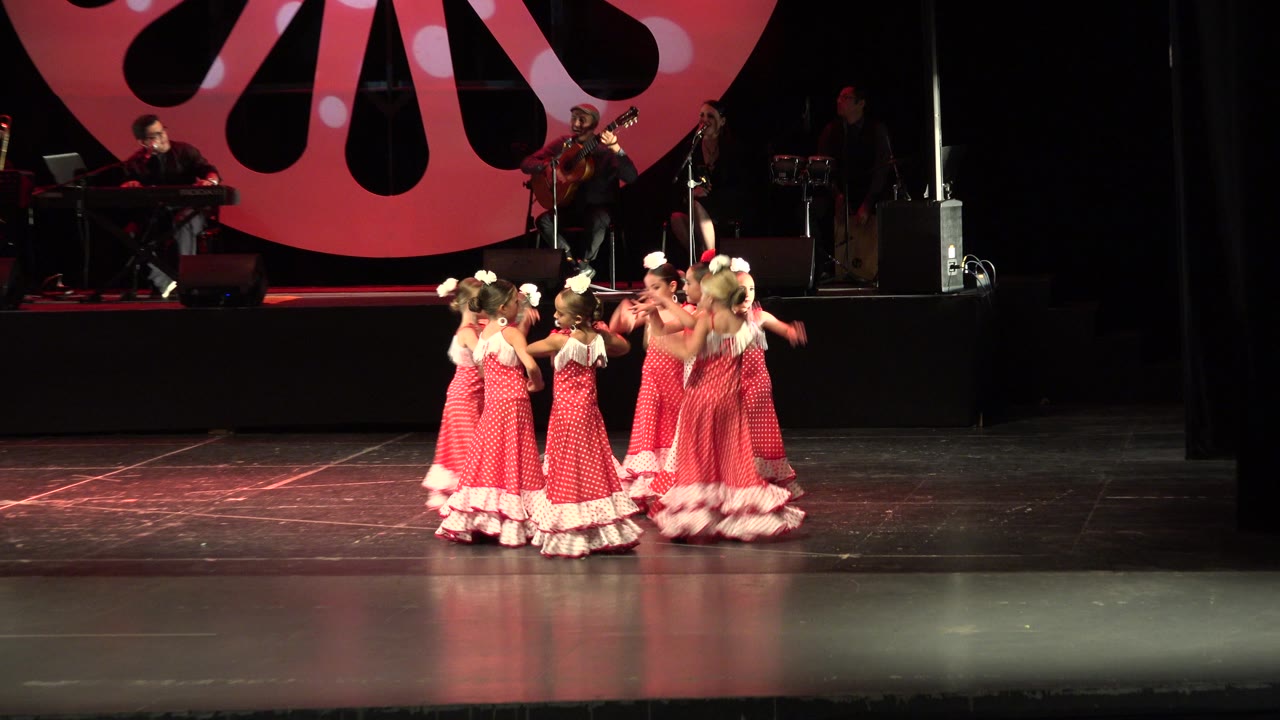 The group of girls in red and white dresses moves toward stage right in Cabo San Lucas, Mexico, with the live band and circular red stage graphic still visible behind them.