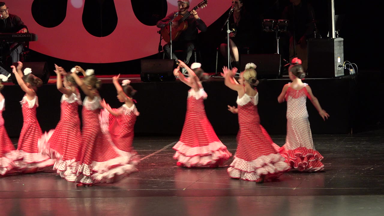 Girls in red and white dresses dance on a stage with musicians behind them.