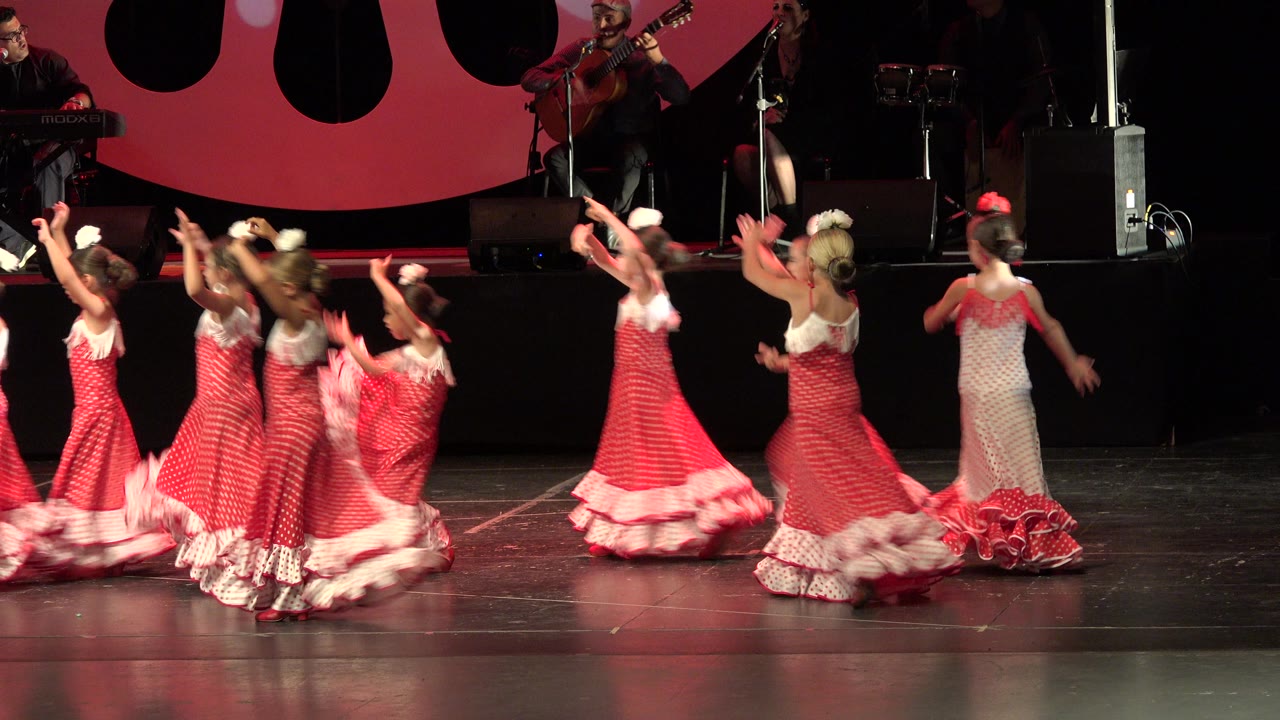 Children in matching red and white dance costumes move through the center of the Cabo San Lucas stage while musicians remain in the dark background.