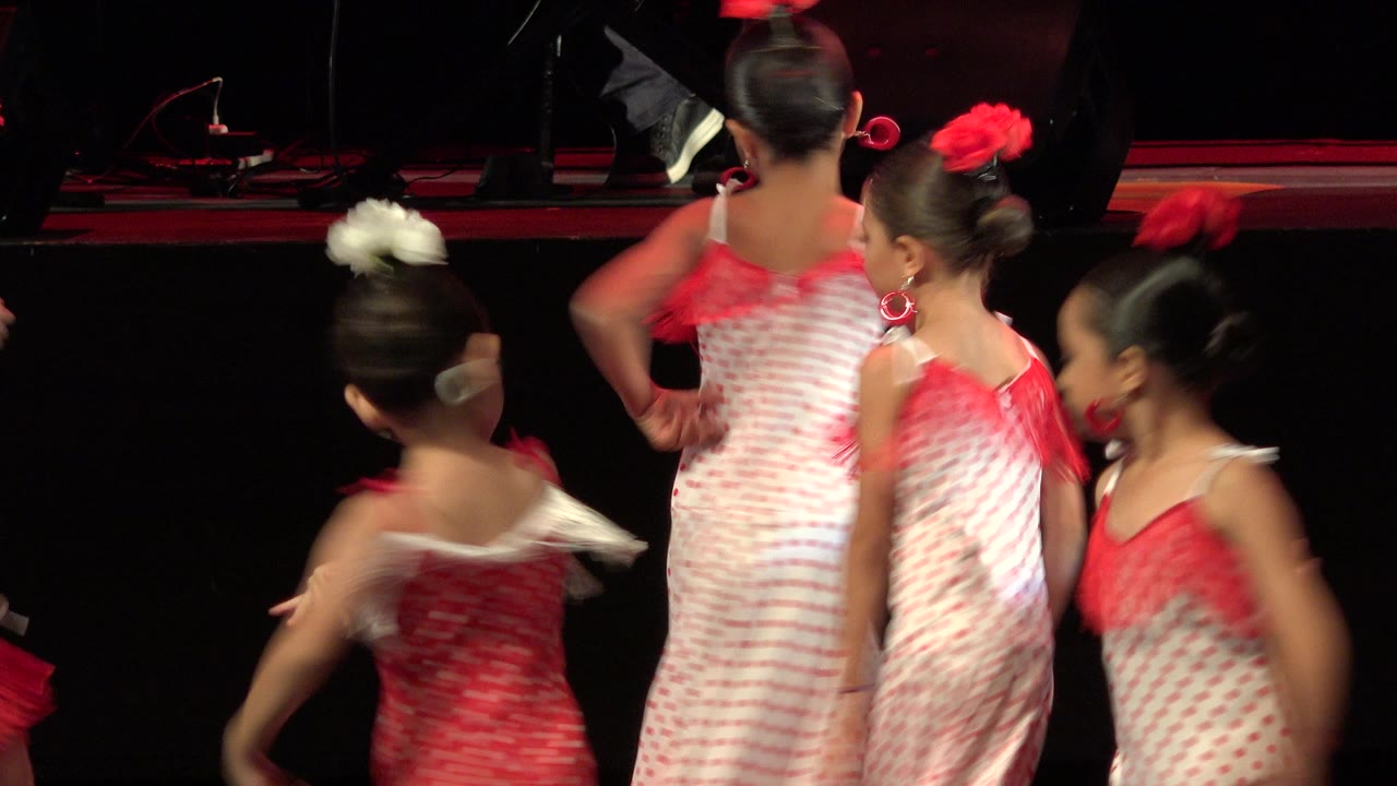 Girls in red and white recital dresses crossing on a dark stage.