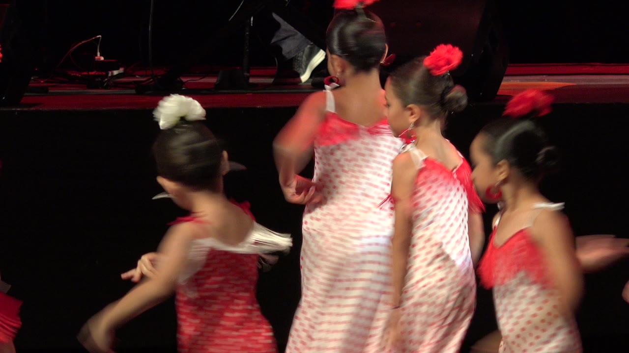 Several young girls in red and white dresses turn and move together on the stage, with dark background equipment and red stage light visible.