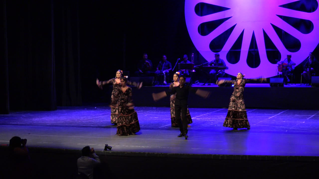 Five young dancers perform on a blue-lit stage with musicians behind them.