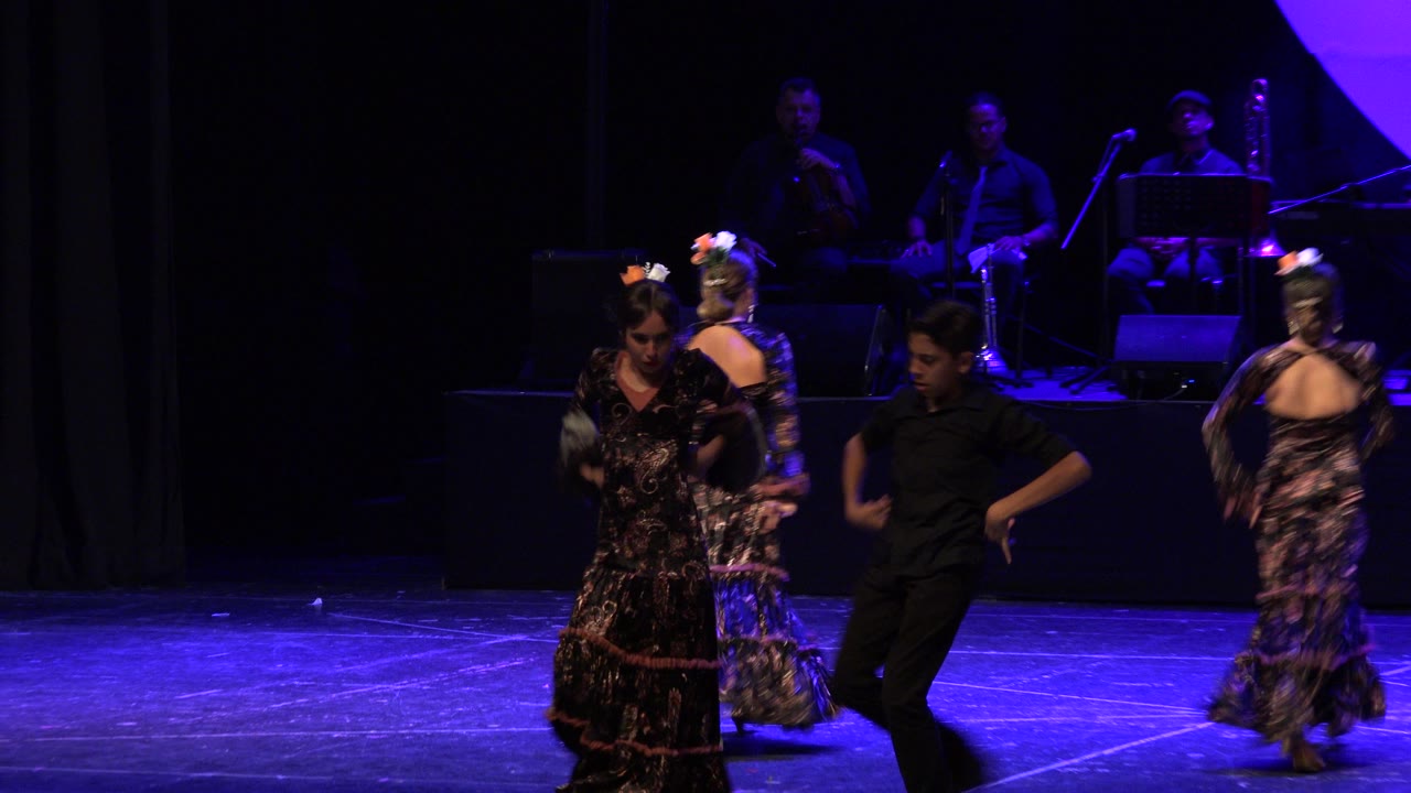 Young dancers perform on a blue-lit stage with musicians in the background.