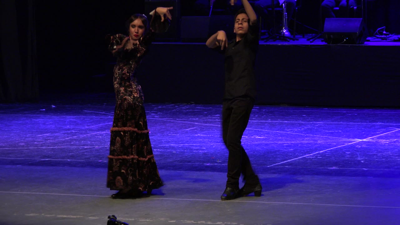Two adult dancers stand on a blue-lit stage with raised arms.