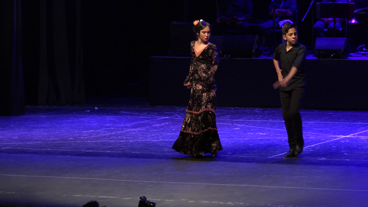 Two visible performers stand apart on a blue-lit stage in Cabo San Lucas, Mexico, with a dark band area behind them at the start of the clip.