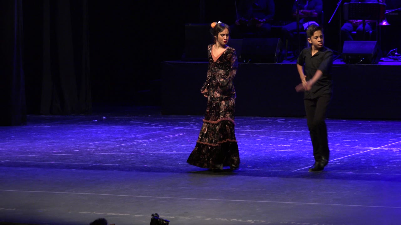 The performers hold poised positions on the blue-lit Cabo San Lucas stage, with the dancer in black standing to the right and the patterned dress visible at left.
