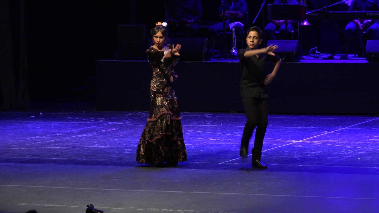 The Cabo San Lucas dance performance continues with the woman and man holding similar forward hand shapes under saturated blue stage light.