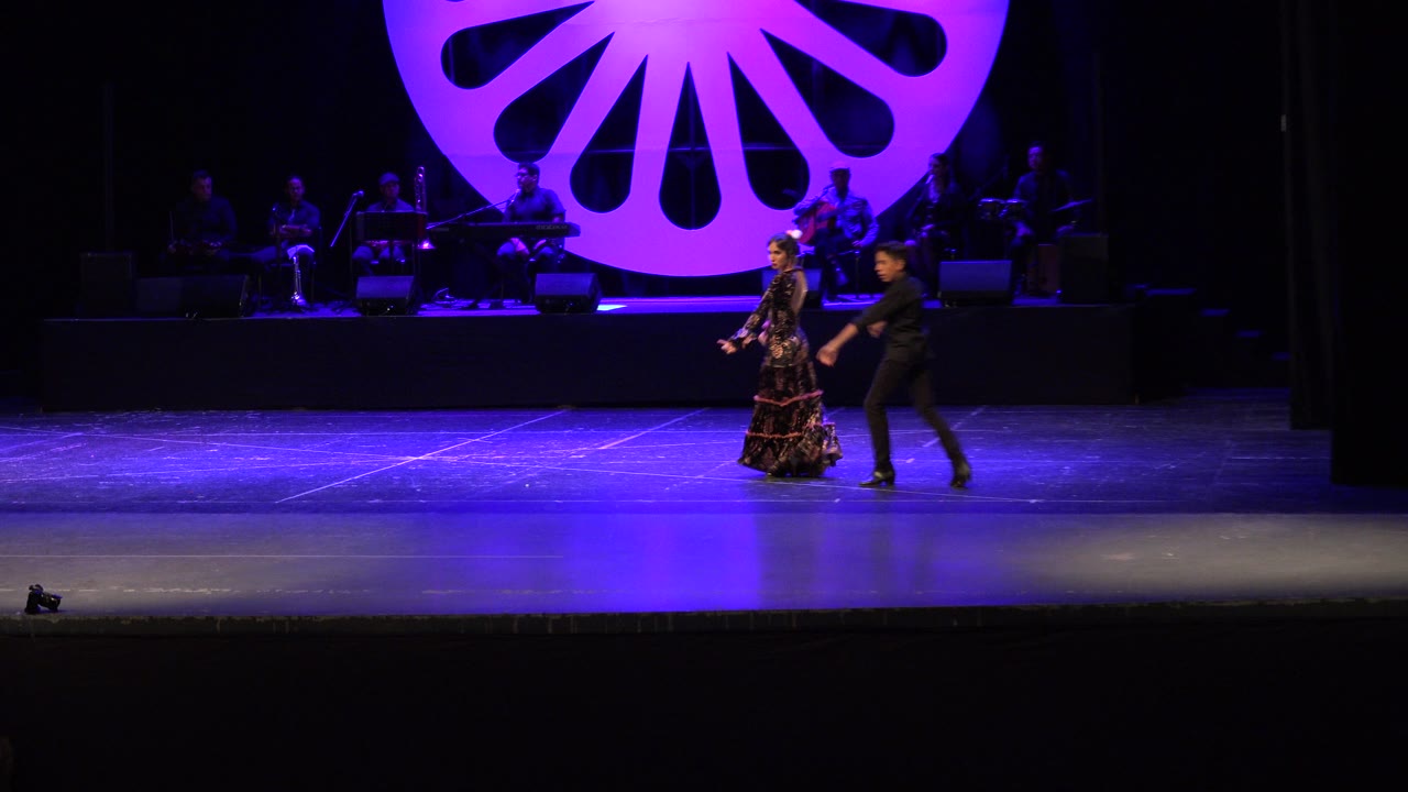 Two dancers cross a purple-lit stage with musicians behind them.