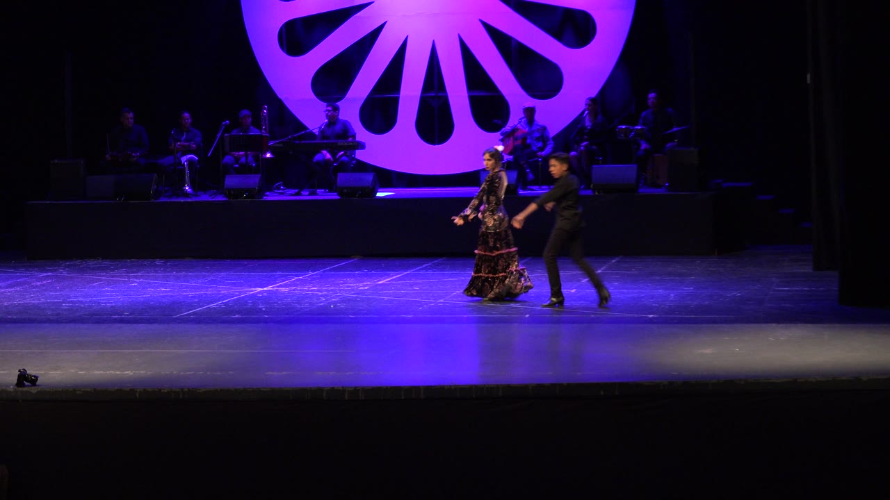 The dancers continue across the center of the Cabo San Lucas stage while the band remains dimly visible behind the purple scenic backdrop.