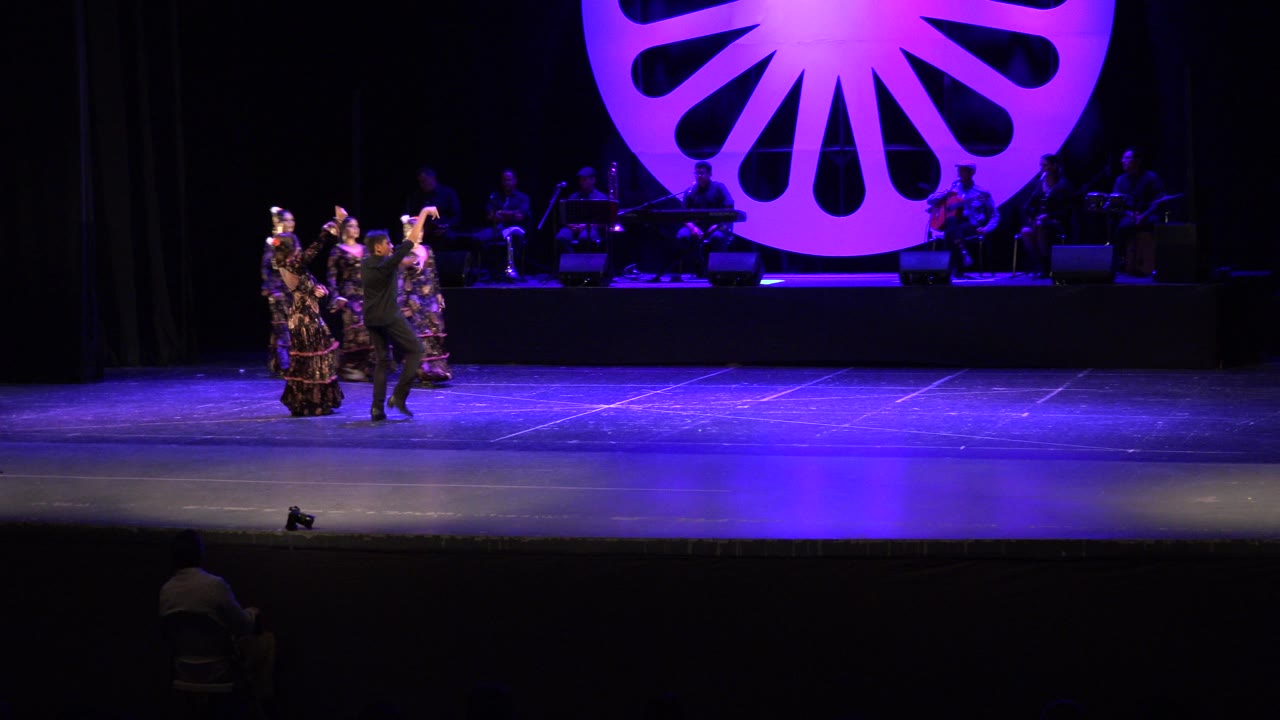 Dancers on a purple-lit stage with musicians and a fan-shaped backdrop.