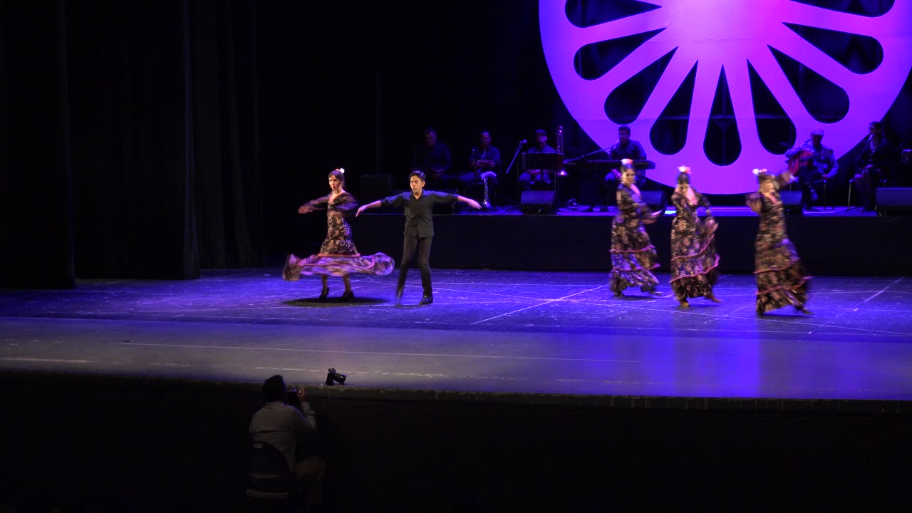 In Cabo San Lucas, dancers in dresses and a dark suit perform across a purple-lit theater stage while musicians sit in shadow behind a large circular backdrop.