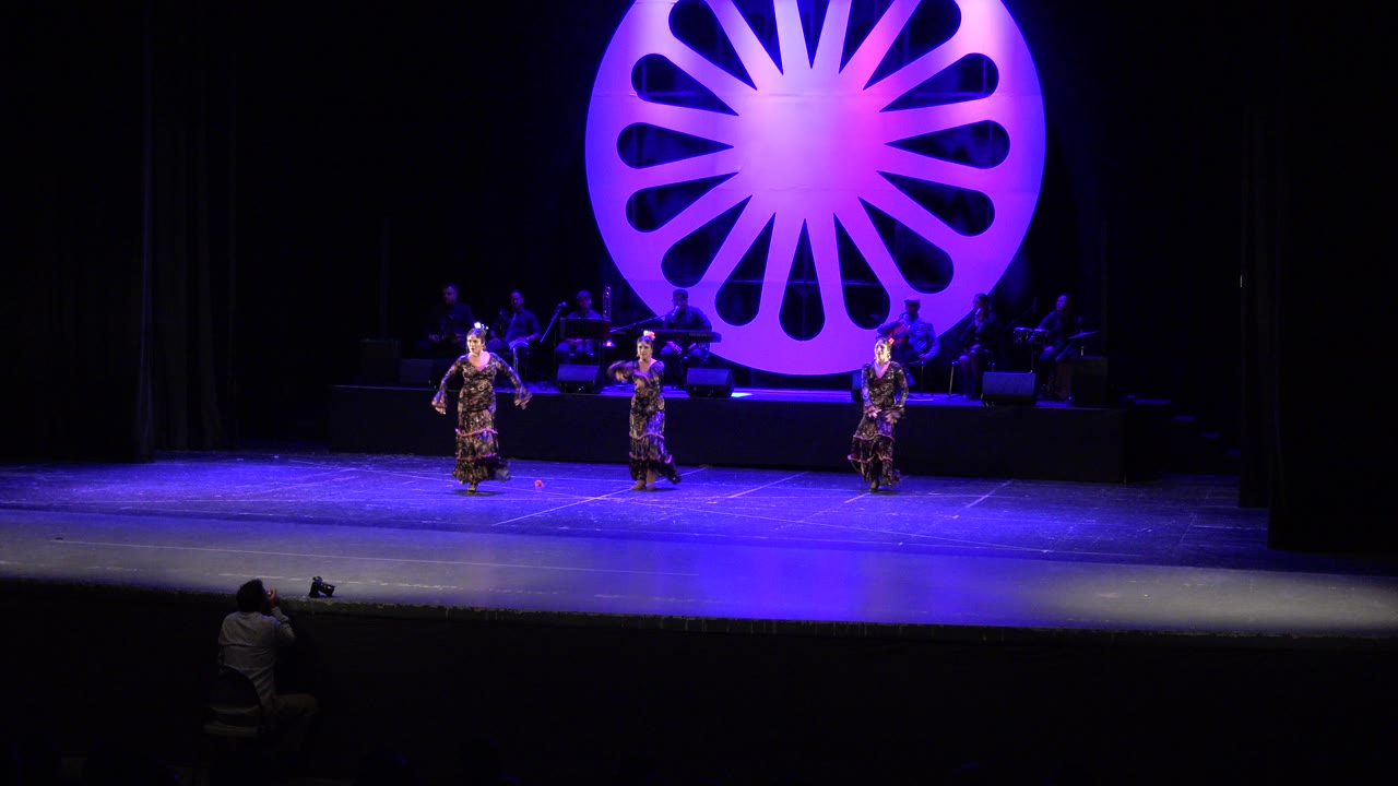 Three women dance on a blue-lit stage with a purple circular backdrop.
