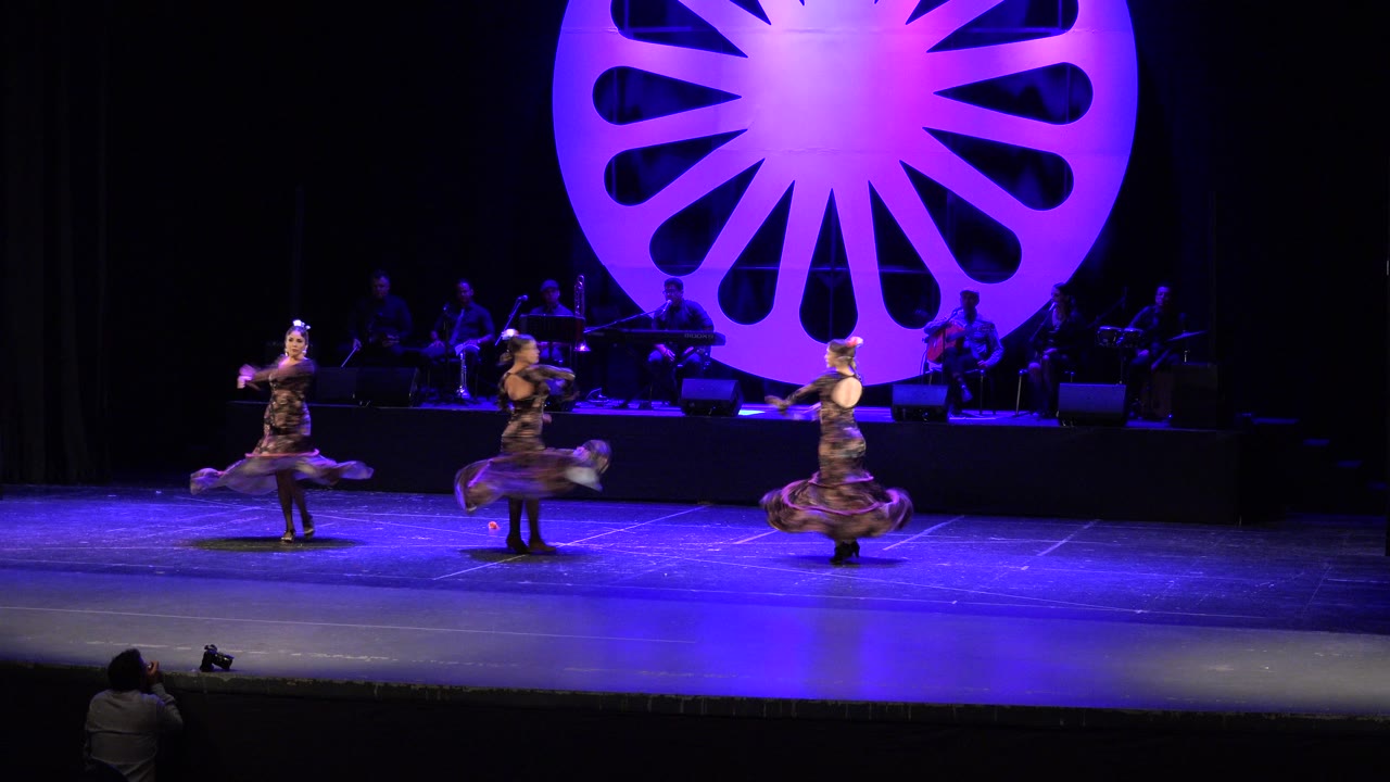 Three women dance on a purple-lit stage with musicians behind them.
