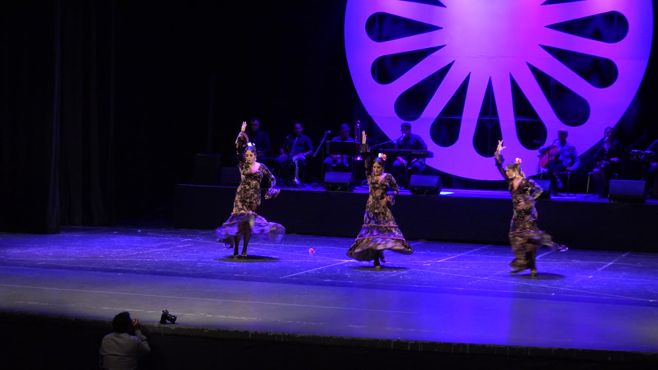 Three women dance on a blue lit stage with musicians behind them.
