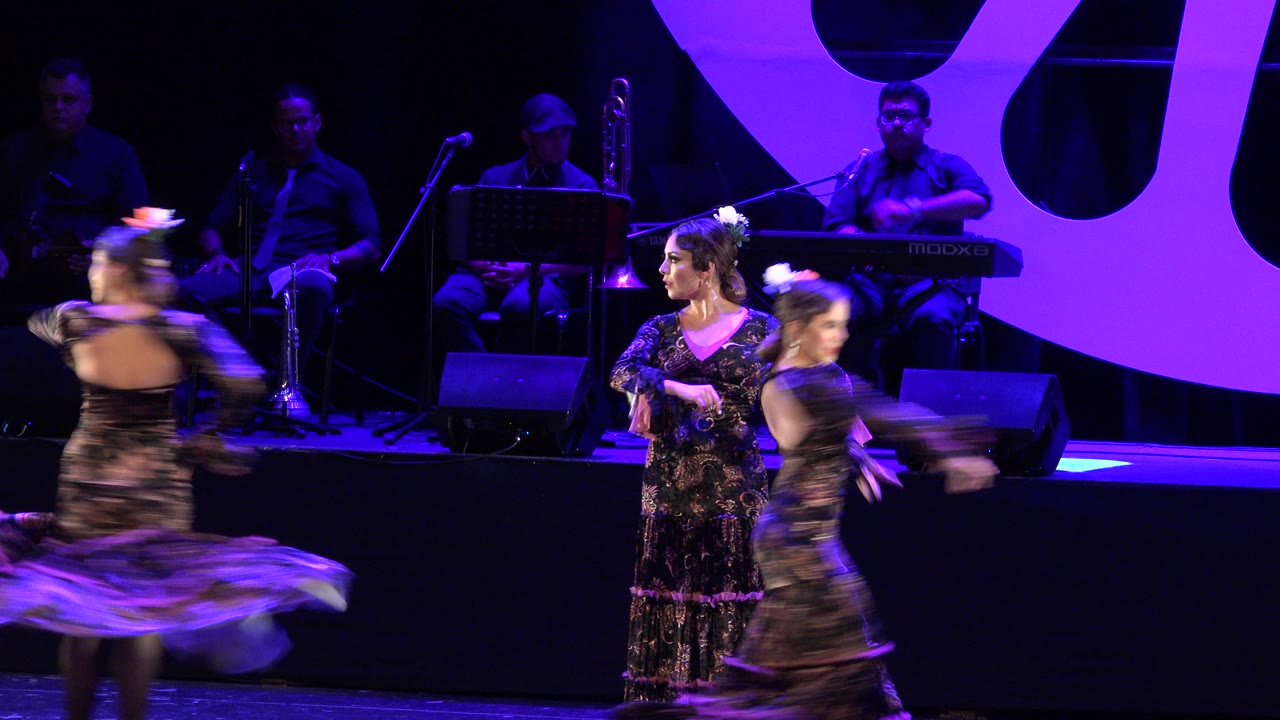 In Cabo San Lucas, Mexico, women in dark floral dresses move across a purple-lit indoor stage while seated musicians and microphone stands sit in the background.
