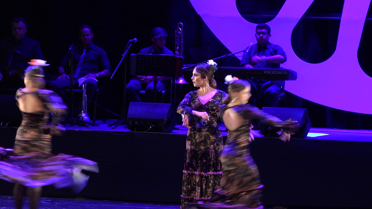 In Cabo San Lucas, Mexico, two dancers blur through the foreground while another woman stands near center stage under purple lighting with musicians seated behind her.