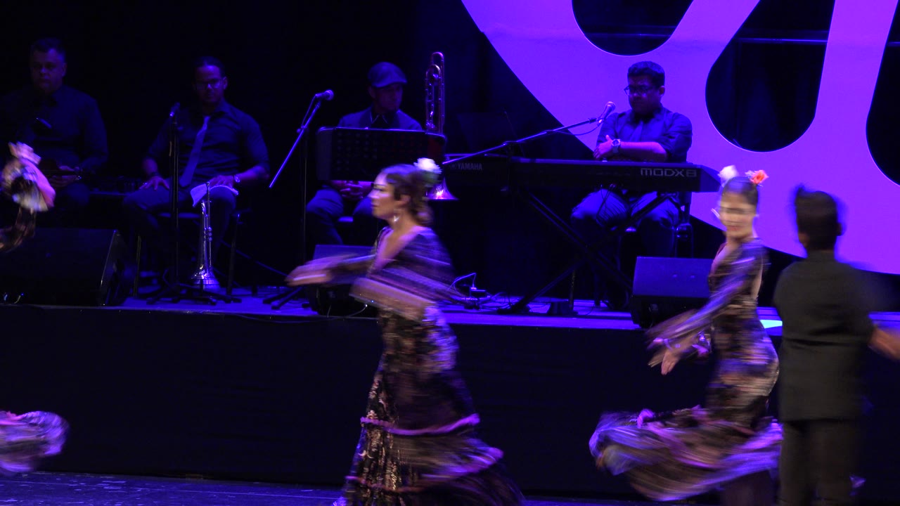 Dancers move across the stage while a man in dark clothing appears at right.