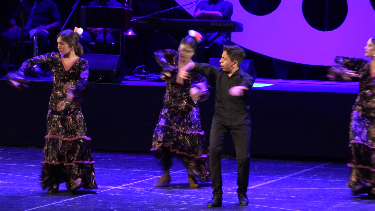 Young dancers continue the routine in Cabo San Lucas, Mexico, with one girl turning at center and a boy in black stepping beside her under purple-blue light.