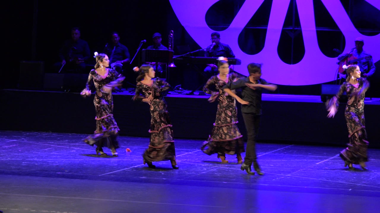 In Cabo San Lucas, Mexico, dancers in dark patterned costumes perform across a blue-purple stage while musicians sit in shadow behind a large purple set piece.
