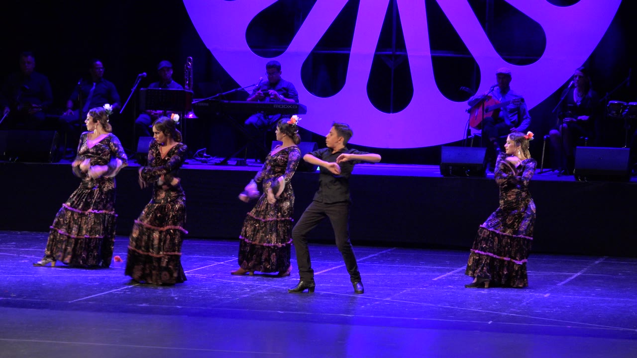 In Cabo San Lucas, Mexico, the male dancer in black extends an arm while women in ruffled floral dresses hold positions across the blue stage beneath purple theatrical lighting.