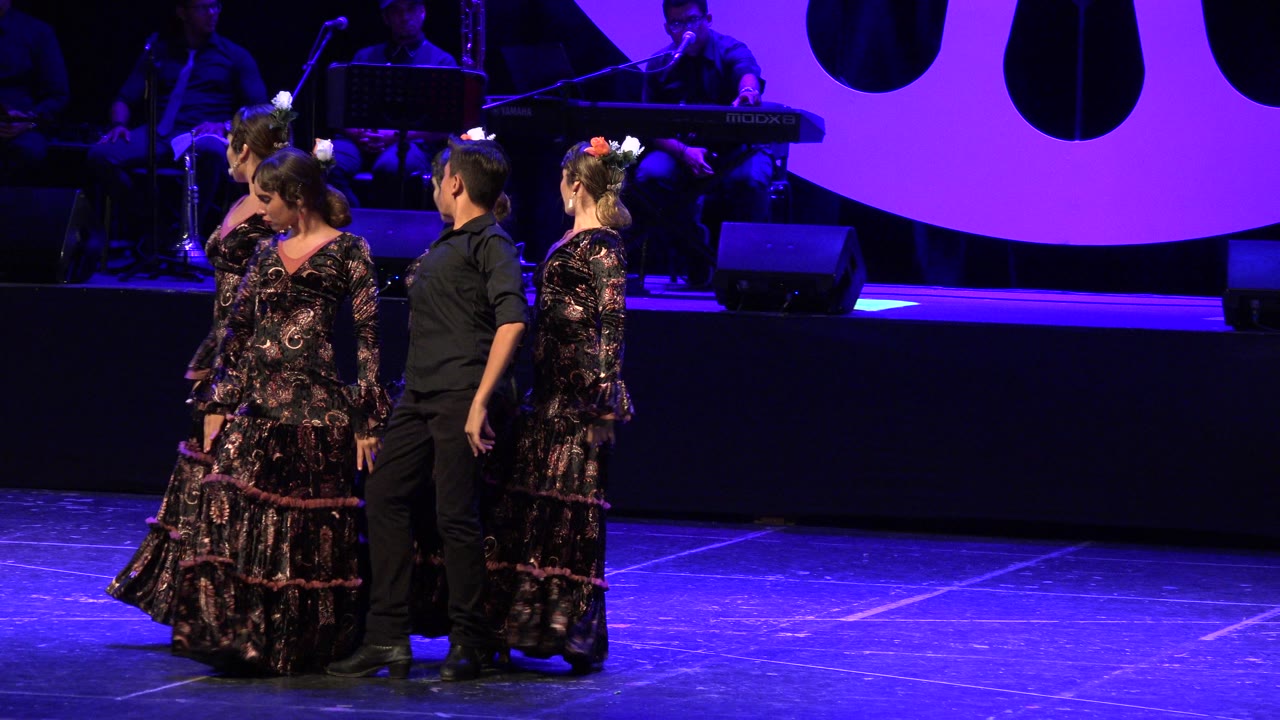 Four dancers perform on a purple-lit stage in Cabo San Lucas, with three women in patterned ruffled dresses, one male dancer in black, and shadowed musicians behind them.