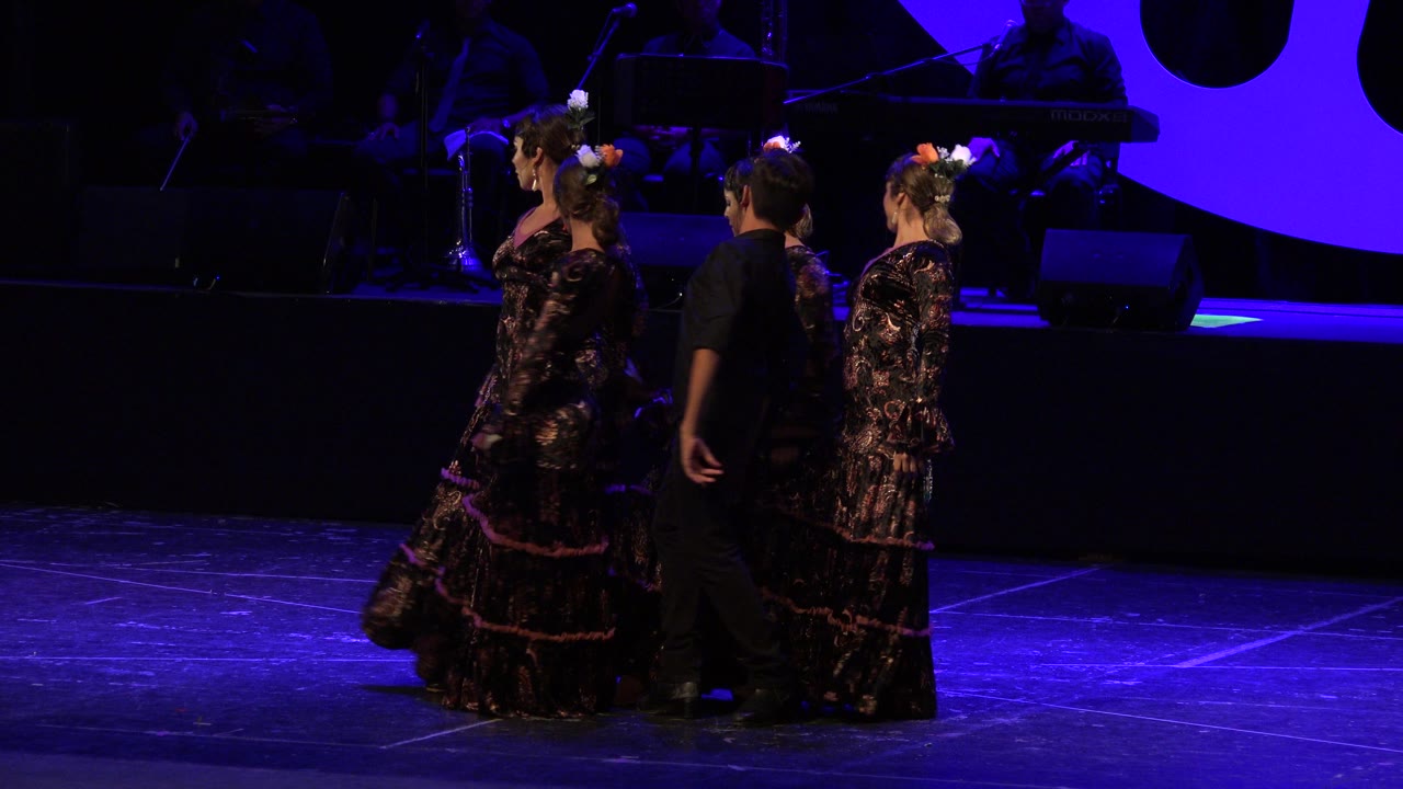Three dancers in Cabo San Lucas, Mexico stand near the left side of a dark stage, with floral dresses, hair flowers, and a man in black visible under blue-purple light.