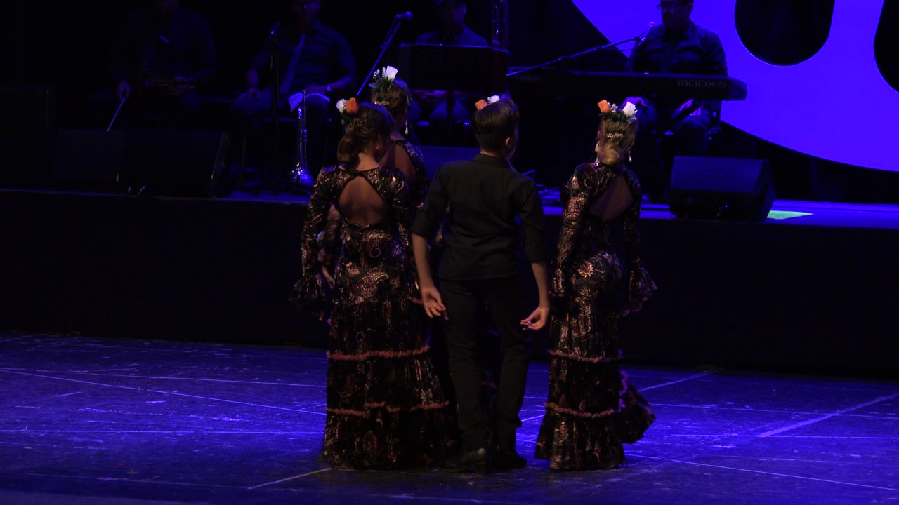 Three performers face away from the camera in Cabo San Lucas, Mexico, centered against a blue-lit backdrop with dark musicians and instruments behind them.