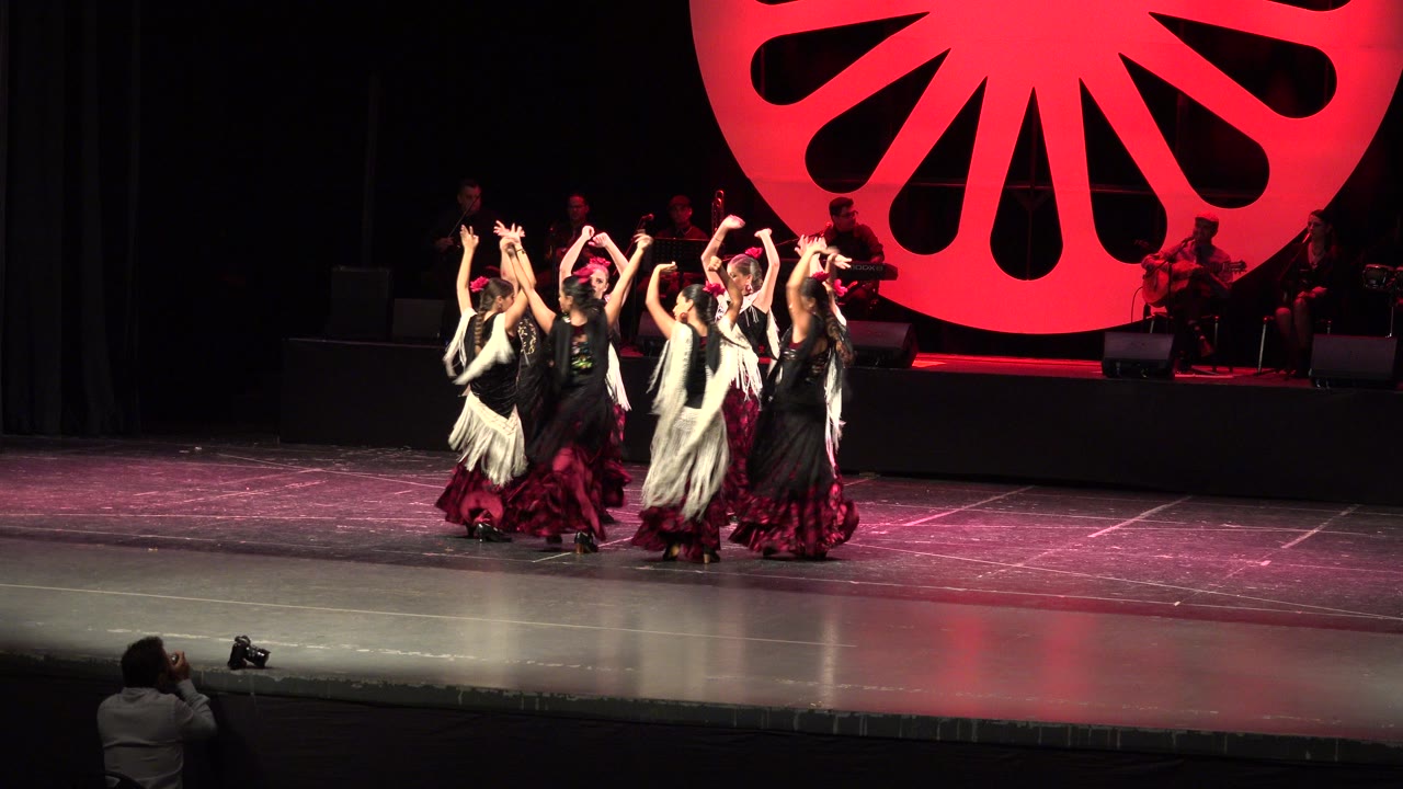 Young female dancers in black and burgundy costumes huddle with raised arms on a stage in Cabo San Lucas, Mexico, with a large red backdrop and a photographer at the front of the audience area.