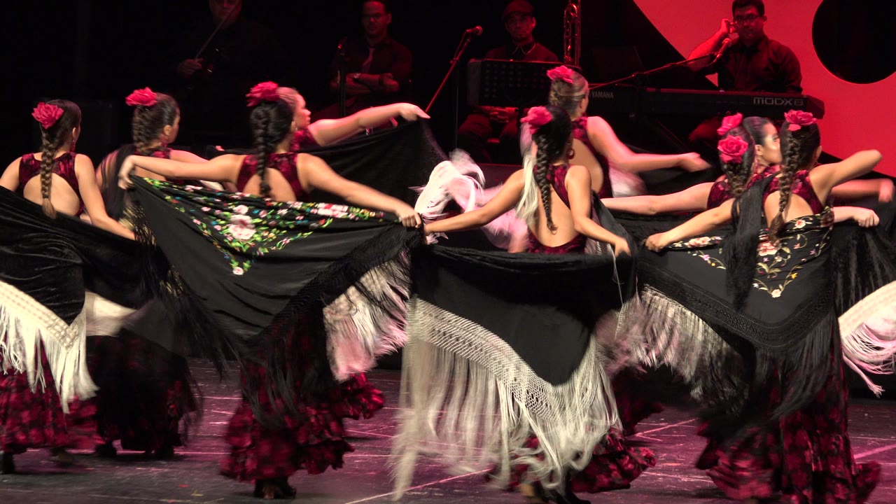 Girls in black shawls with fringe dance on a lit stage.
