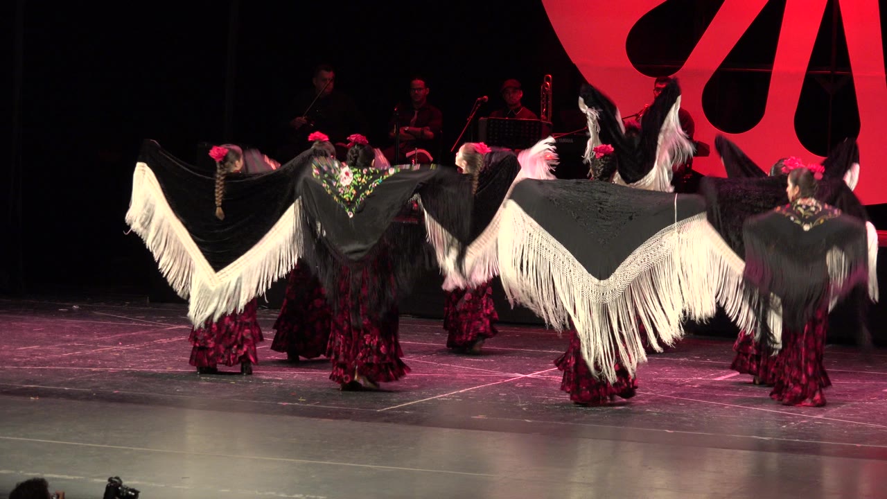 Dancers in red dresses hold black shawls with white fringe on a theater stage.