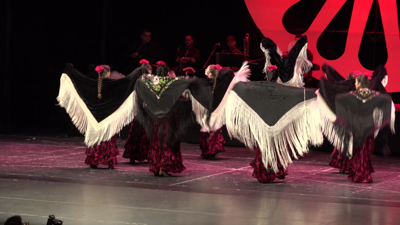 Dancers in Cabo San Lucas keep their black shawls stretched outward, with white fringe forming a wide line in front of the seated orchestra.