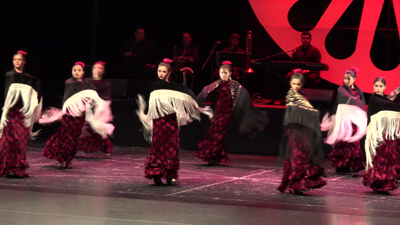 Dancers in Cabo San Lucas rotate through the stage formation as white fringed shawls blur in motion and red lighting outlines the musicians behind them.
