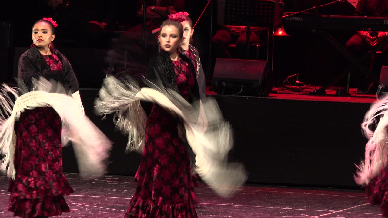 Women in red dresses dance with fringed shawls on a dark stage.
