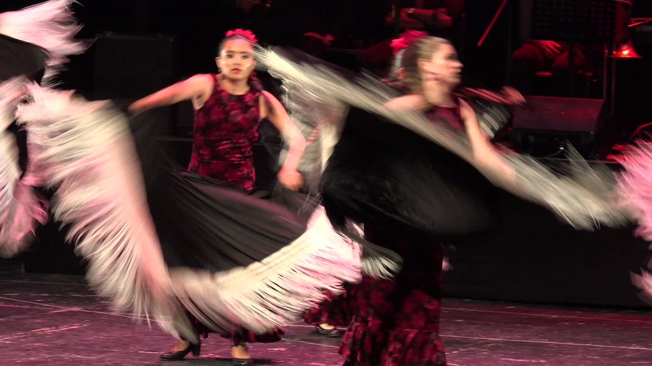 At the start of the Cabo San Lucas performance, a young dancer in a dark red floral dress faces forward while black and white fringed fabric blurs across the stage.