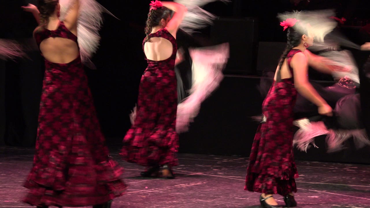 Woman in a red and black dress turns with a pale fringed shawl on a dark stage.