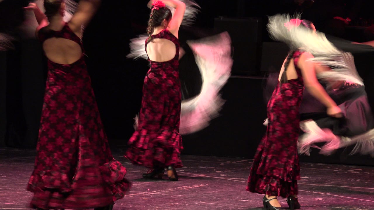 In Cabo San Lucas, two women in matching red and black dresses perform on a dark stage while pale fringed shawls blur across the frame.