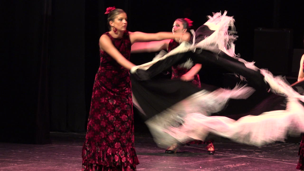 A front dancer in a red-and-black dress reaches across the stage while another dancer behind her moves a black scarf with pale fringe in Cabo San Lucas, Mexico.
