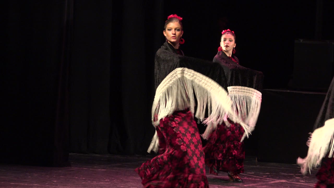 Two female dancers perform on a dark stage in Cabo San Lucas, Mexico, with black shawls, red patterned dresses, and white fringe sweeping across the foreground.