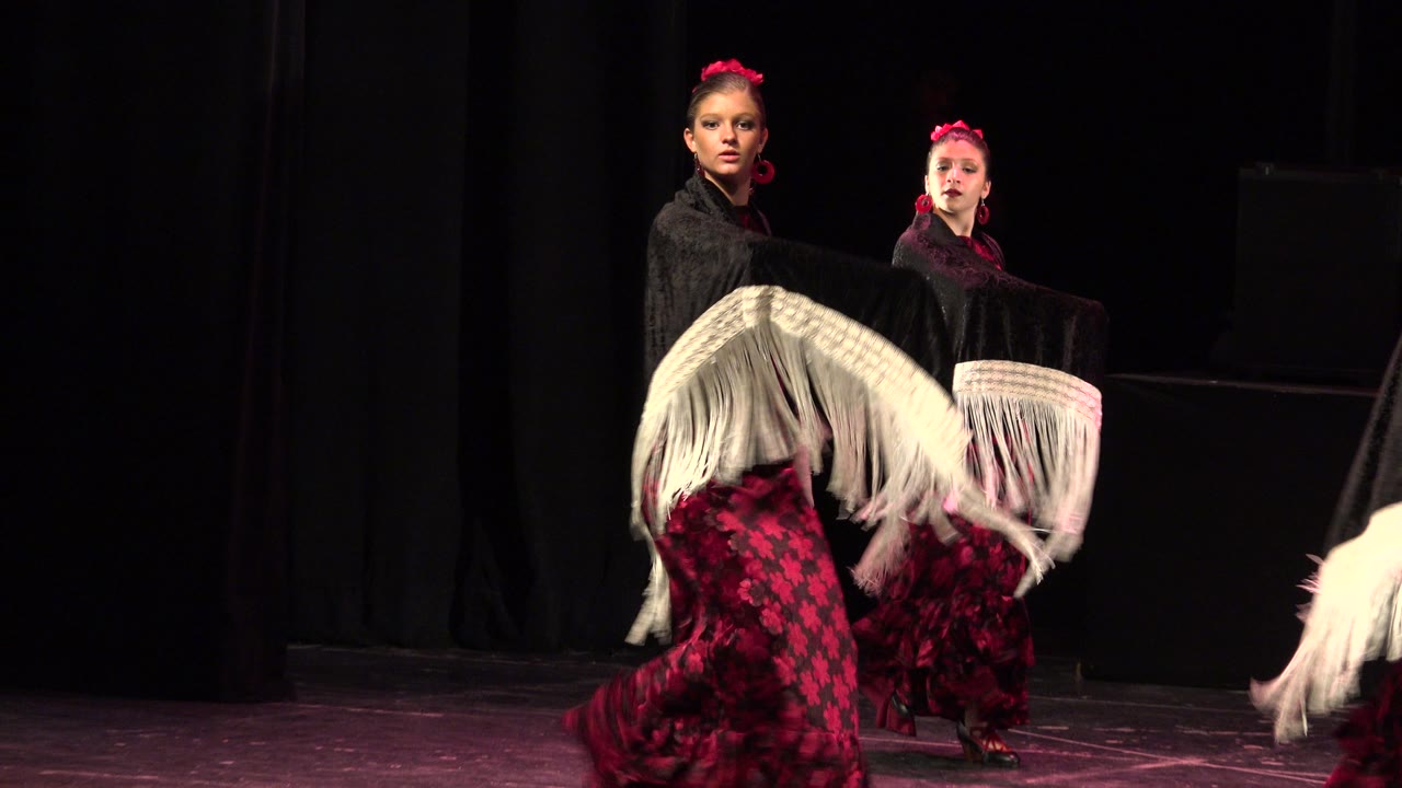 Two dancers remain centered on the Cabo San Lucas stage, their faces lit while layered white fringe crosses the lower foreground.
