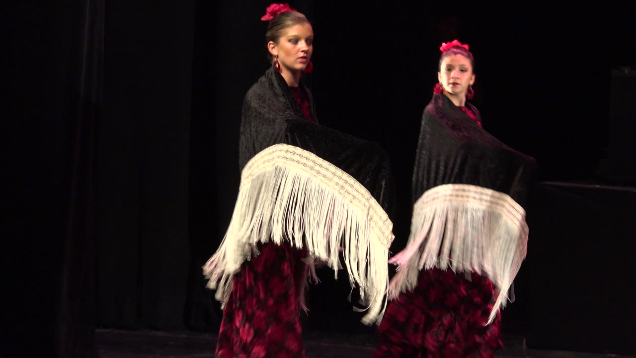 Two dancers in black shawls turn beside white fringe fabric.