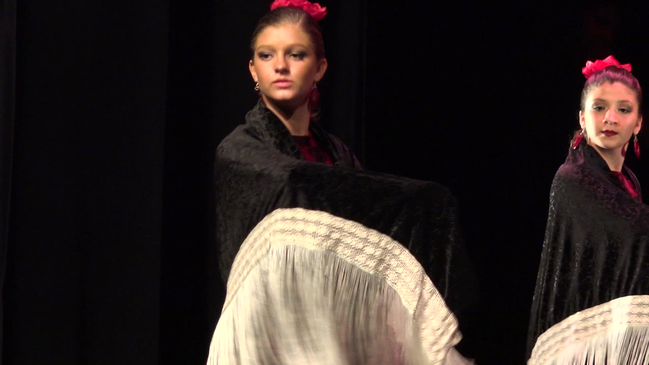 In Cabo San Lucas, two young dancers stand under warm stage light, one close to camera holding a black shawl over white fringe while another appears farther right.
