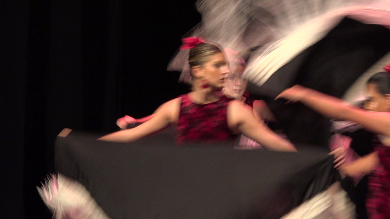 Dancers in Cabo San Lucas continue the routine as a white fringed prop blurs above and a wide black fabric panel fills the lower foreground.